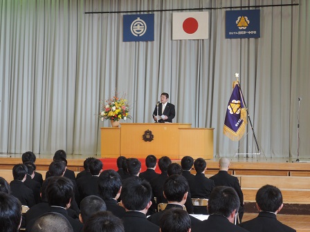 校旗が置かれた卒業式のステージ上の演台で、祝辞を述べている市長と 座って聞いている卒業生たちの写真