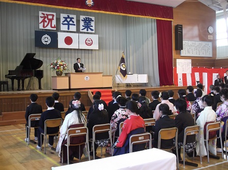 祝卒業と書かれたの幕が張られ、校旗が置かれたステージ上で、 祝辞を述べている市長と座って聞いているスーツ姿や袴姿の卒業生たちの写真