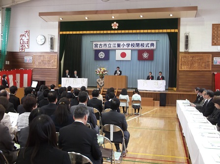 宮古市立江繋小学校閉校式典の横断幕が張られたステージ上で、来賓の方々が同席する中、 あいさつをしている市長と座って聞いている在校生を含む多くの参加者の方々の写真