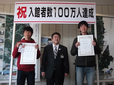 祝入館者数100万人達成の横断幕の下で、それぞれ認定証を持った水野翼さんと橋本晶さんの間に市長が立って撮影した写真