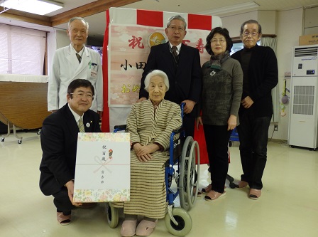 車椅子に乗った小田切マキさんの横で、市長が100歳の記念品を持ち、 その後ろに小田切さんの関係者4人が立って撮影した写真