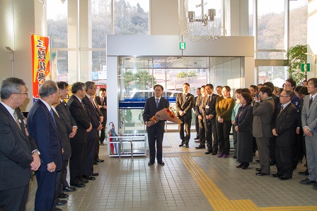 副市長を退任される名越一郎さんが花束を持ち、市役所入口付近で通路両脇に立っている多くの市職員に見送られる写真