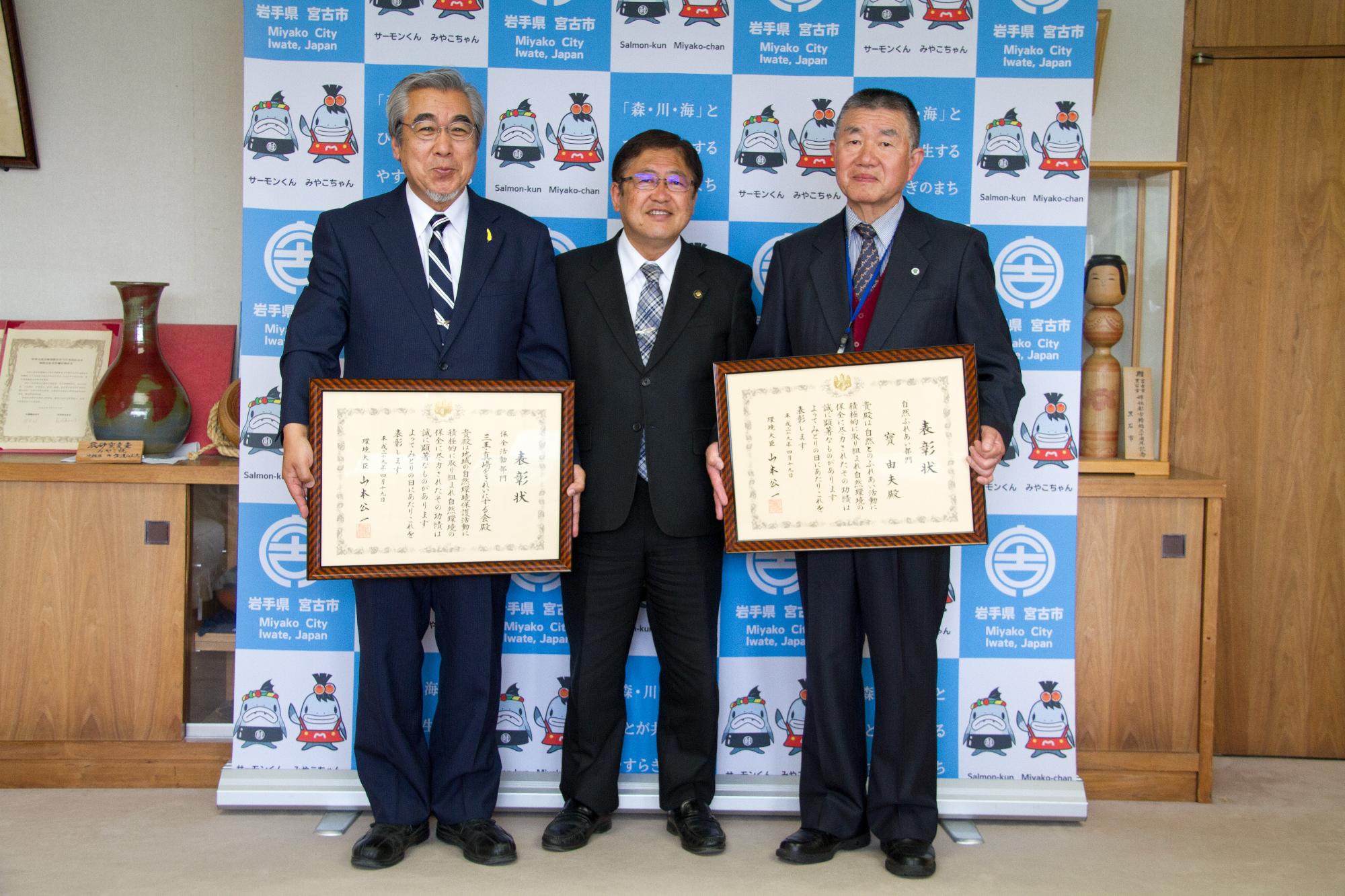 バックパネルの前で、市長を中心に賞状を持つ男性2名が両側に立っている記念写真