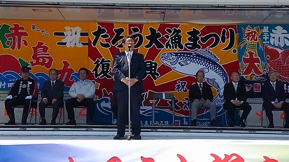 大漁旗の前のパイプ椅子に座る6名の関係者と、スタンドマイクに向かい話をする市長の写真