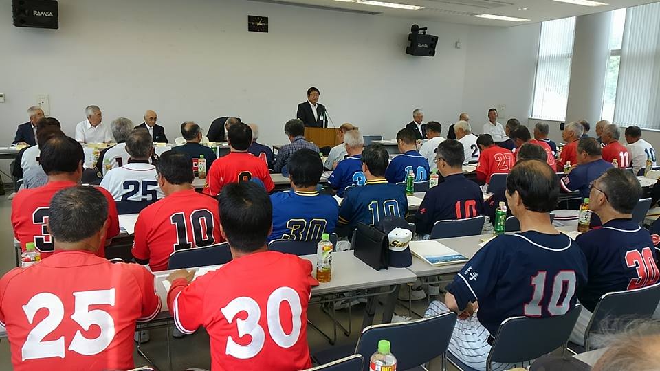 演台の前に立つ市長と、野球のユニフォームを着た選手たちが、部屋に並べられたテーブルの席についている写真