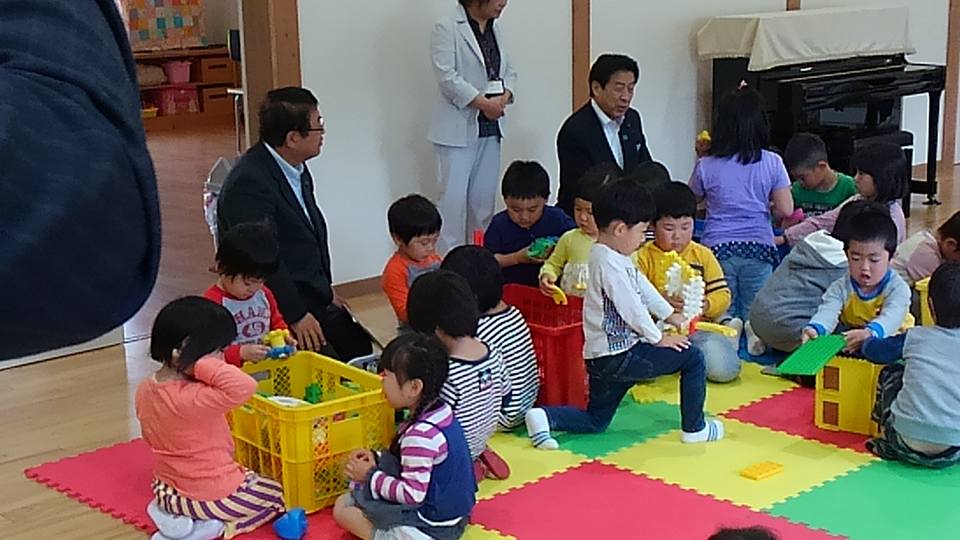 赤色、黄色、緑色などのジョイントマットが敷かれた部屋で、おもちゃで遊ぶ子供たちと、子供たちを見ている様子の市長と男性の写真