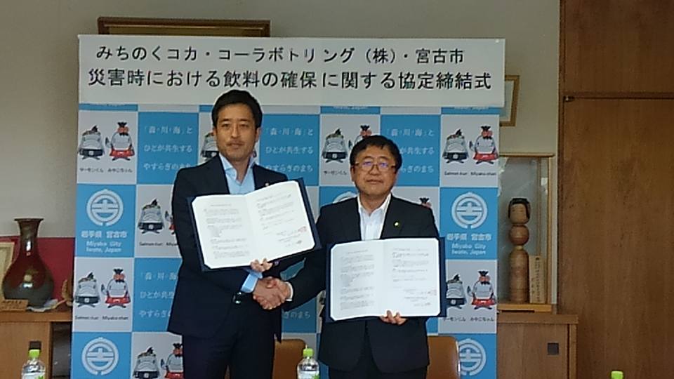 「みちのくコカ・コーラボトリング株式会社・宮古市災害時における飲料の確保に関する協定締結式」と書かれた紙が貼られたバックパネルの前で、締結書を左手に広げて持ち、右手で握手をする市長と男性の写真
