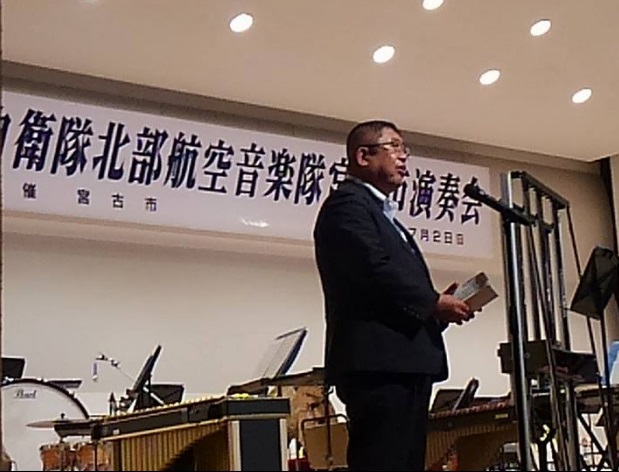 音楽隊の楽器が置かれた会場で、スタンドマイクに向かい話をする市長の写真