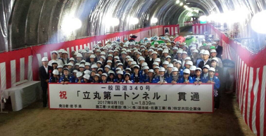 トンネルの壁に紅白幕が貼られ、「祝「立丸第一トンネル」貫通」と書かれた横断幕の後ろにヘルメットを被った児童たちや関係者が並んでいる写真