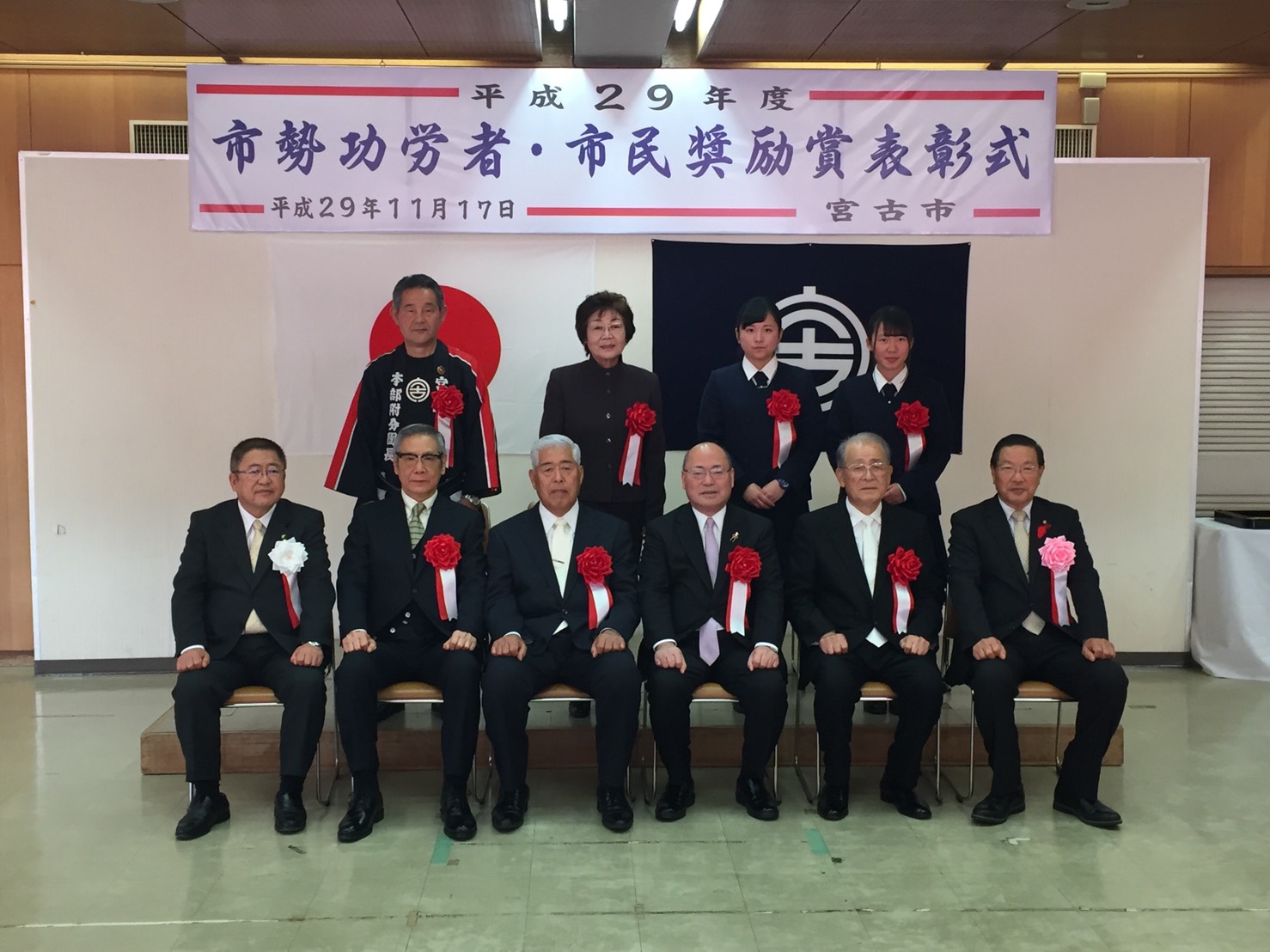 胸章を着けた市長と5名の男性が椅子に座り、後列に立って並ぶ男性1名、女性1名、制服を着た女子学生2名が写った記念写真