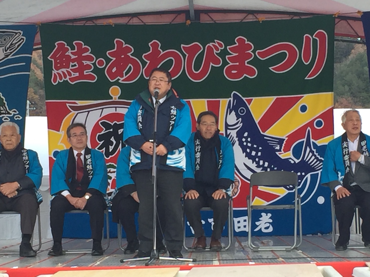 大漁旗が掛けられた舞台で、スタンドマイクに向かい話をする市長の写真