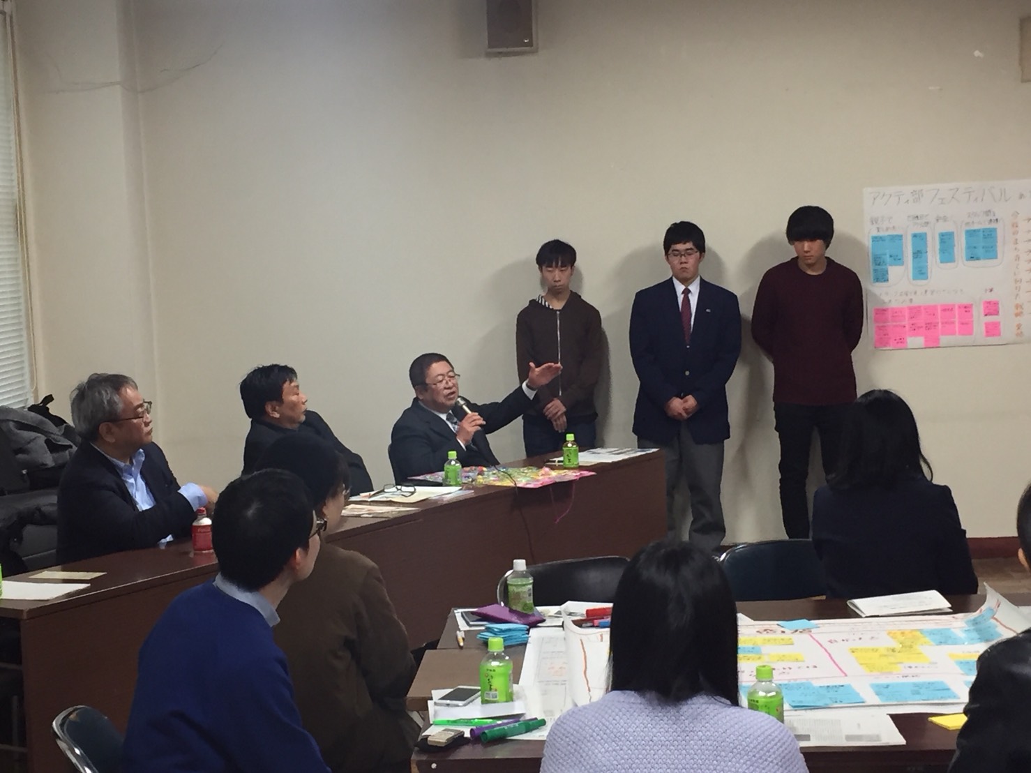 茶色いテーブルの席に着席した2名の男性の隣に座る市長が、右手にマイクを持ち左手を顔の前まであげながら話をしている様子の写真