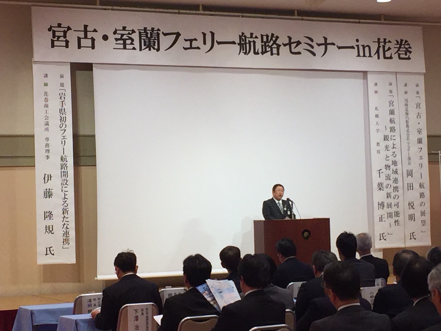 「宮古・室蘭フェリー航路セミナーin花巻」と書かれた横断幕の下の舞台で、演台に置かれたマイクに向かい話をする様子の市長の写真
