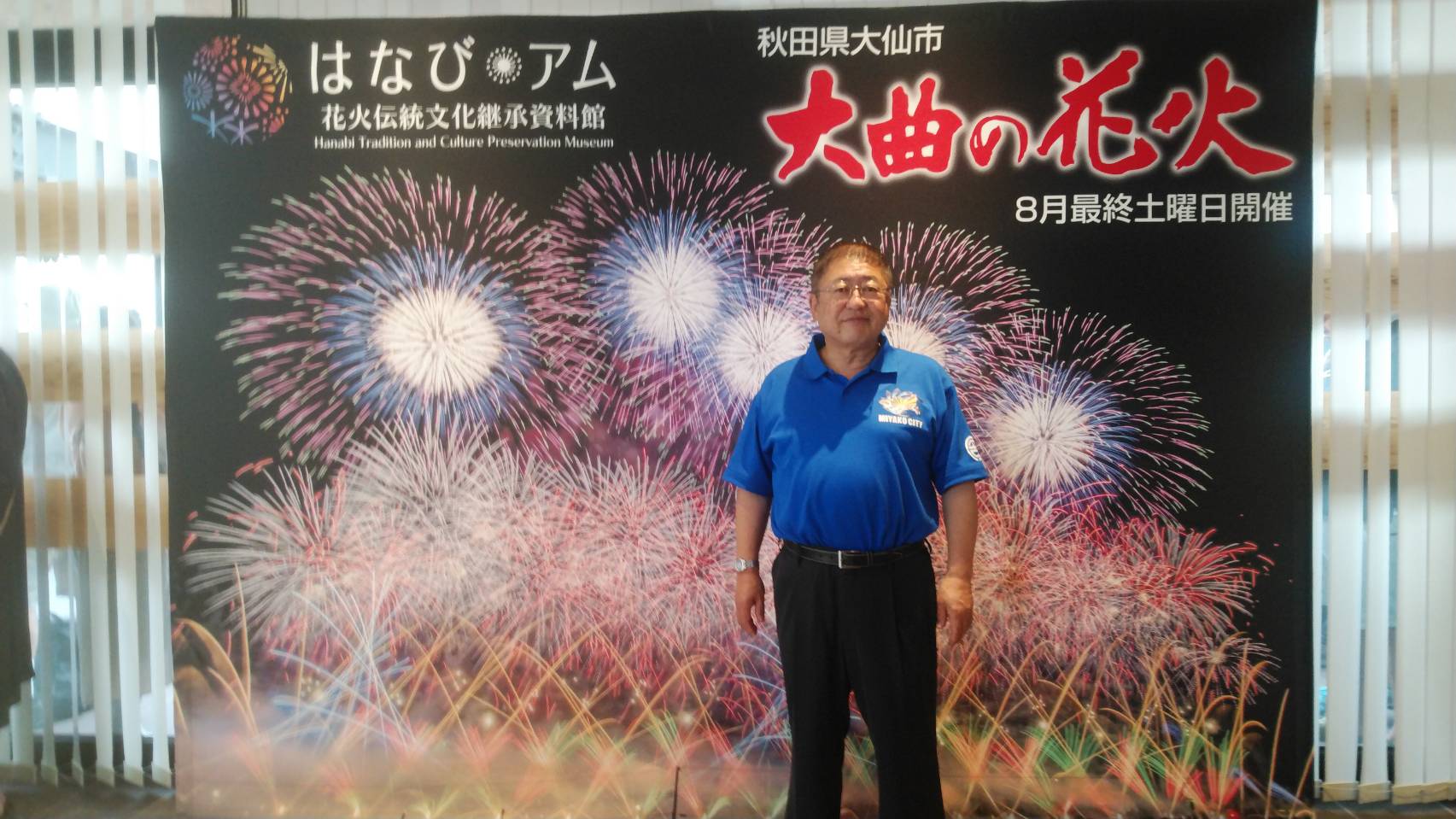 大曲の花火の写真が写った大きなポスターの前に立つ市長を写した記念写真