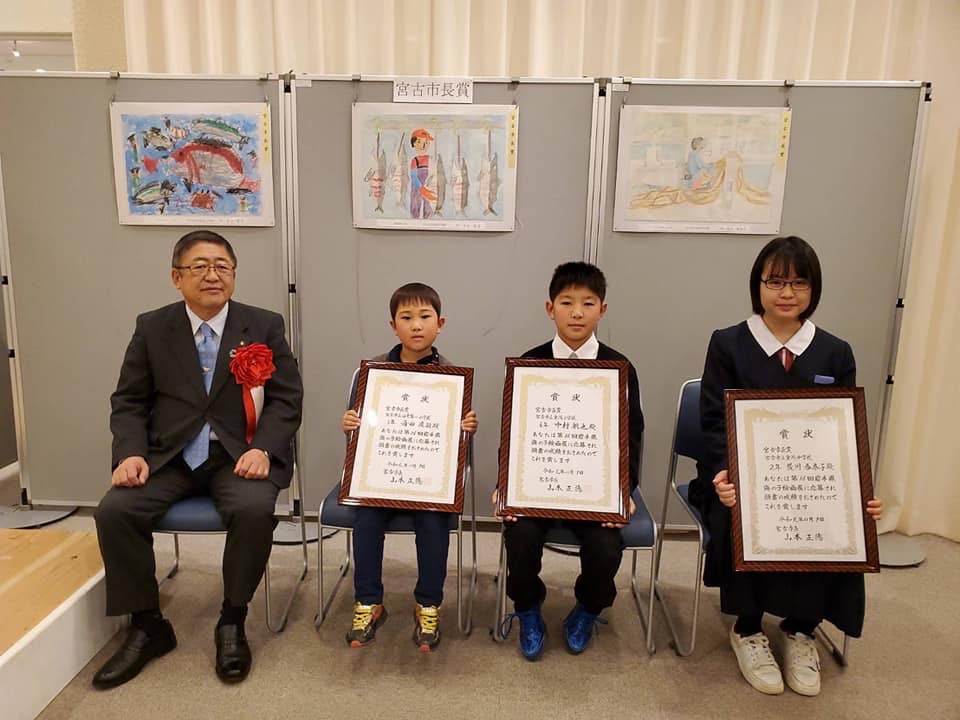 絵が飾られたボードの前で、賞状の額縁を持って椅子に座る男の子2名、女の子1名と市長が写っている記念写真