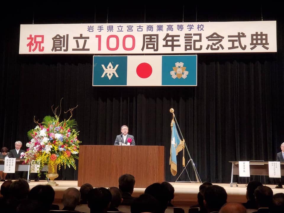 「岩手県立宮古商業高等学校 祝 創立100周年記念式典」と書かれた横断幕や国旗・校旗が吊るされた舞台上で、演台に向か講演をする様子の市長の写真