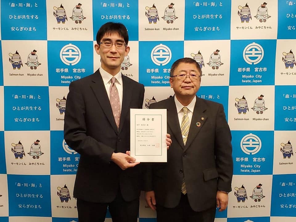 バックパネルの前で、辞令書を両手で持つスーツを着た男性と市長が立って並ぶ記念写真