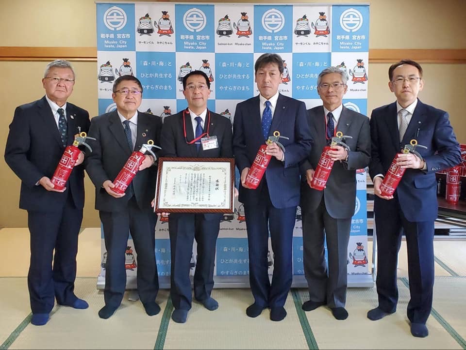 バックパネルの前で消火器を持つ市長を含む5名の男性と賞状の額縁を持つ1名の男性の記念写真
