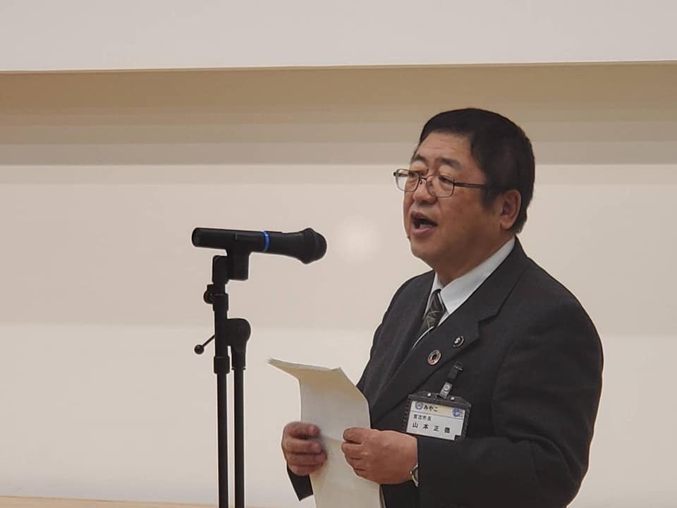 白い用紙を持ってスタンドマイクに向かい話をする様子の市長の写真