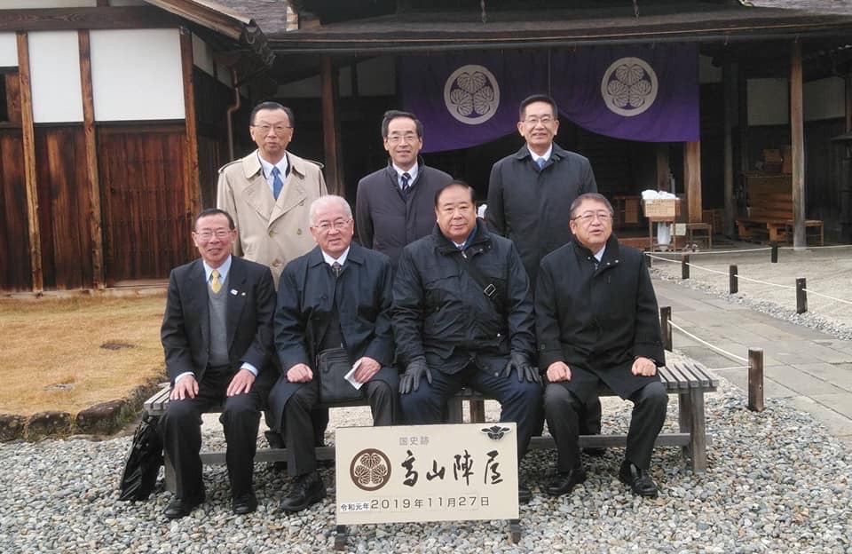 「高山陣屋」と書かれたプレートの後方で、椅子に座る男性3名と市長が前列、直立する男性3名が後列に並んでいる記念写真