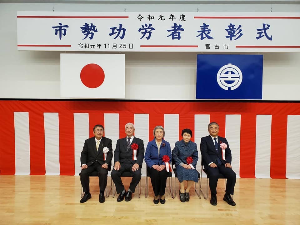 「市勢功労者表彰式」と書かれた横断幕と日本国旗、宮古市の市町村旗が吊るされ、壁に紅白幕が貼られた会場で、椅子に座り横一列に並ぶ市長と功労者の4名の記念写真