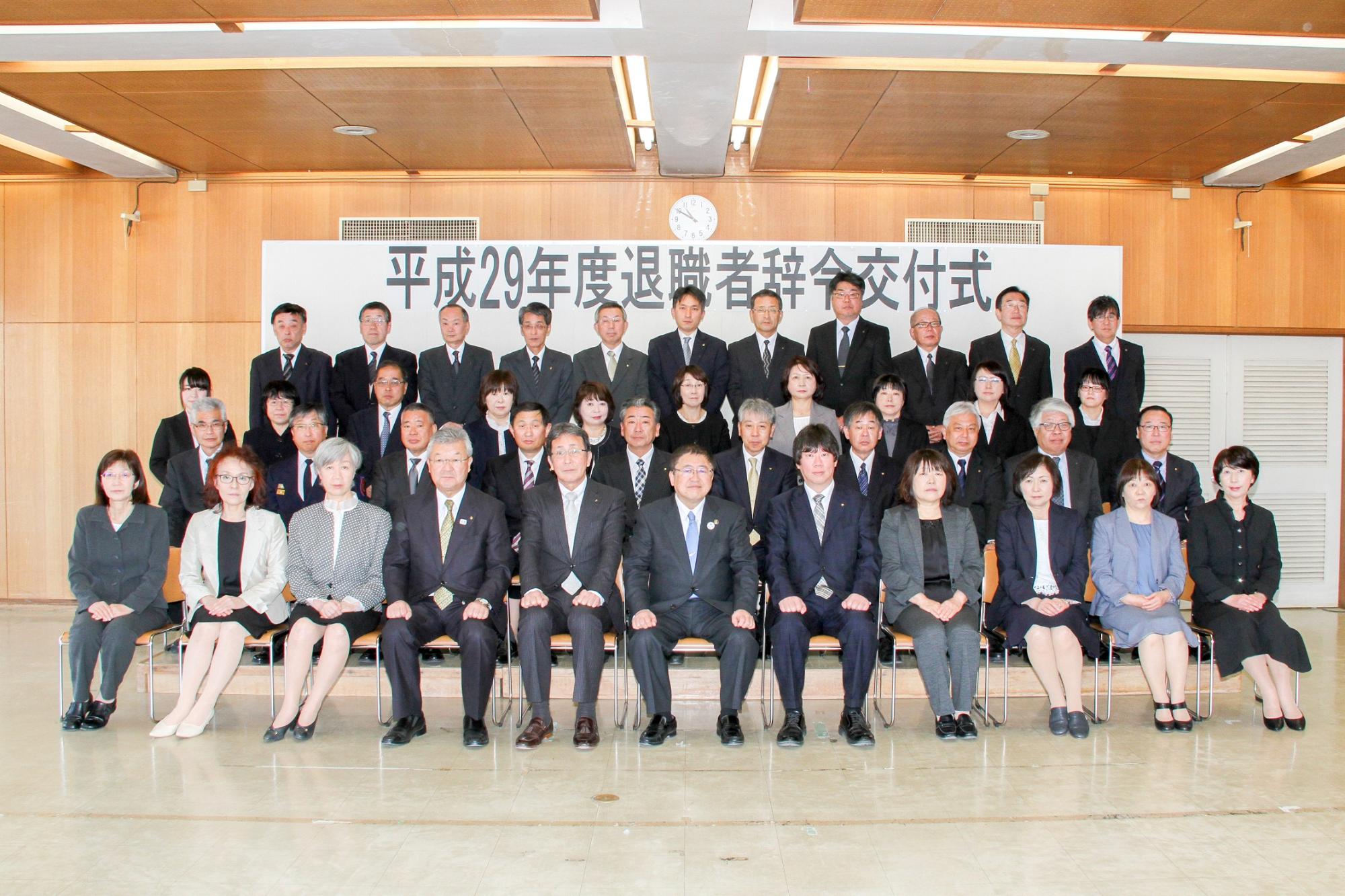 「平成29年度退職者辞令交付式」と書かれたパネルの前で、4列に並ぶ市長と職員の方々の記念写真