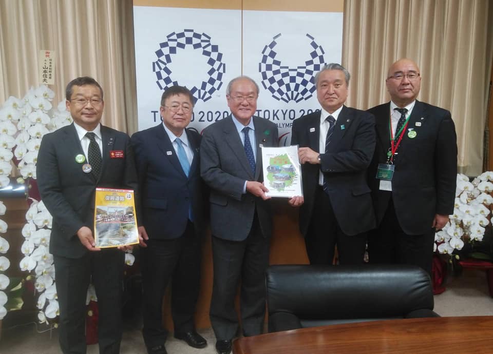 東京オリンピックのエンブレムが描かれたポスターの前で、パンフレットを両手で持つ1名の男性、市長、パンフレットを一緒に持つ男性2名と男性1名が横一列に並んでいる記念写真