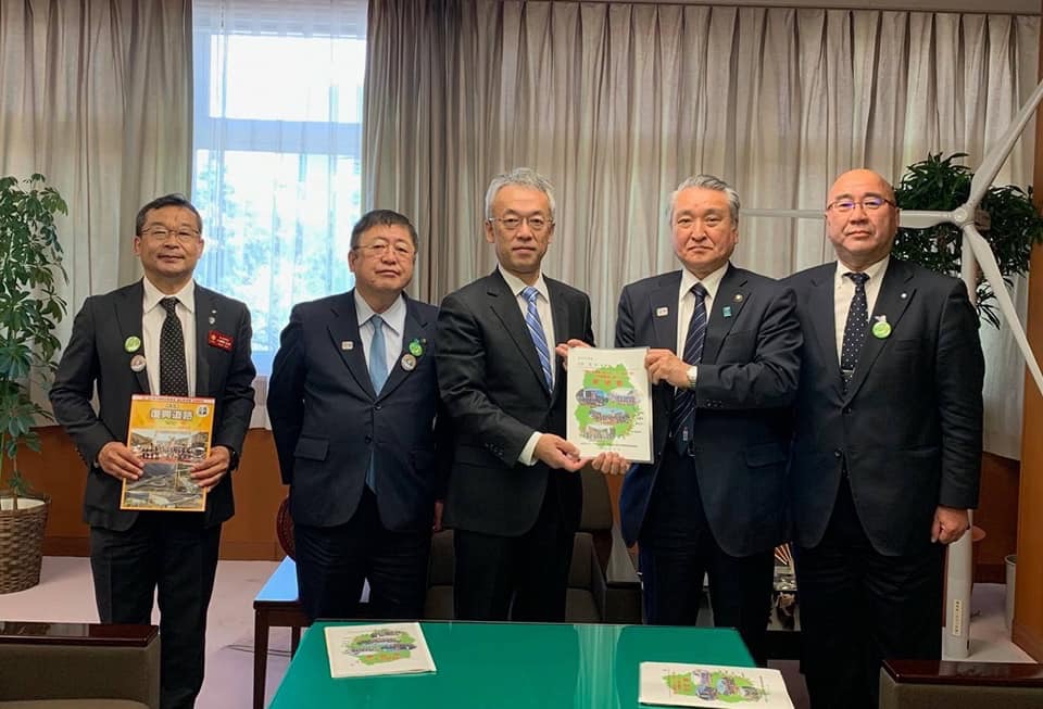 パンフレットを両手で持つ1名の男性、市長、パンフレットを一緒に持つ男性2名と男性1名が横一列に並んでいる記念写真