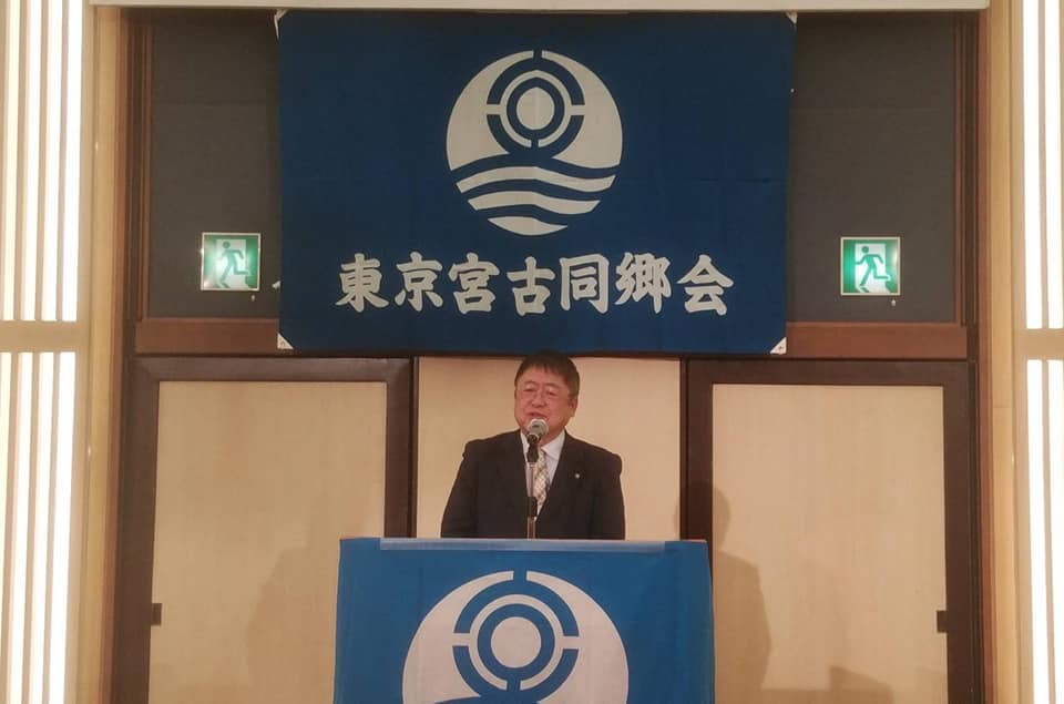 「東京宮古同郷会」と書かれた旗が掲げられた壁の前で、演台に置かれたマイクに向かい話をする様子の市長の写真
