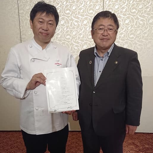 両手でメニュー表を持ちコックコートを着用した奥田政行氏と市長が並んでいる写真