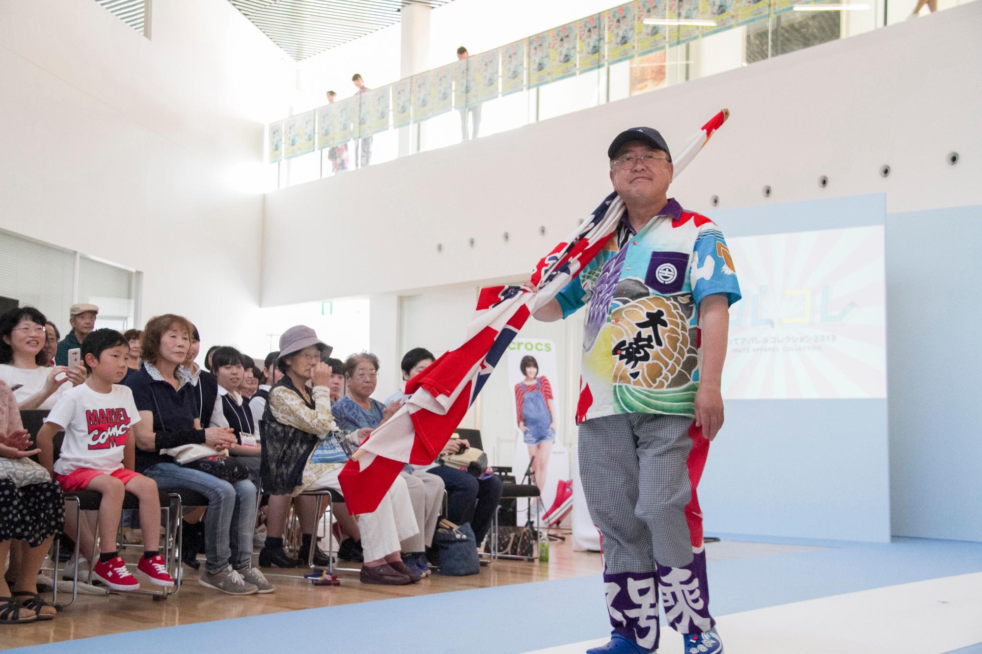 大漁旗を片手に、カラフルな大漁旗のシャツとズボンを着た市長が、観客が座っている前のランウェイを歩いている写真