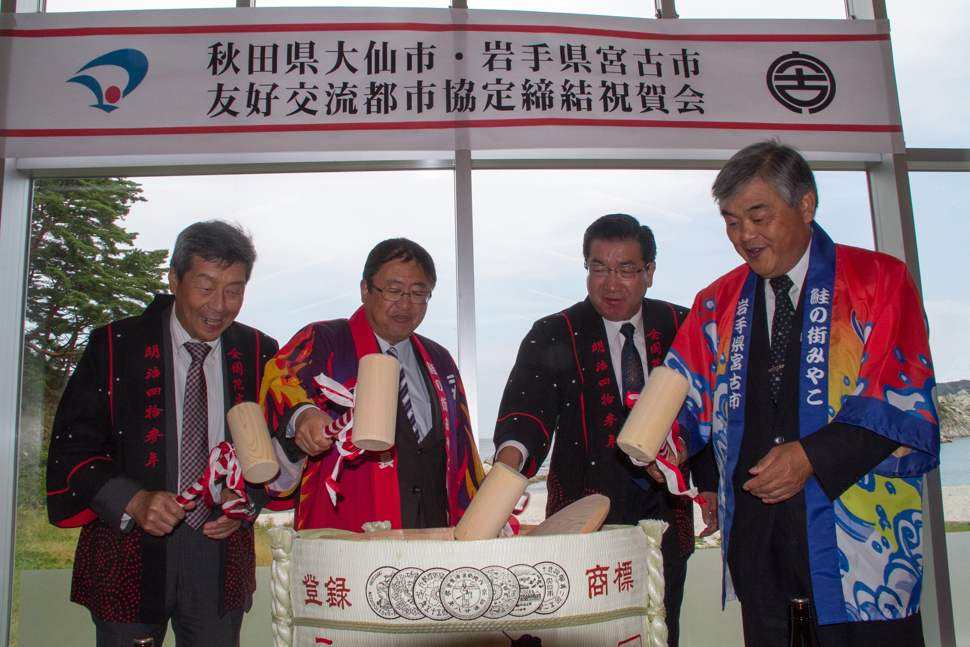 「秋田県大仙市・岩手県宮古市 友好交流都市協定締結祝賀会」と掲げられた横断幕の下、法被を着た市長と関係者が木槌を持ち、鏡開きをしている写真