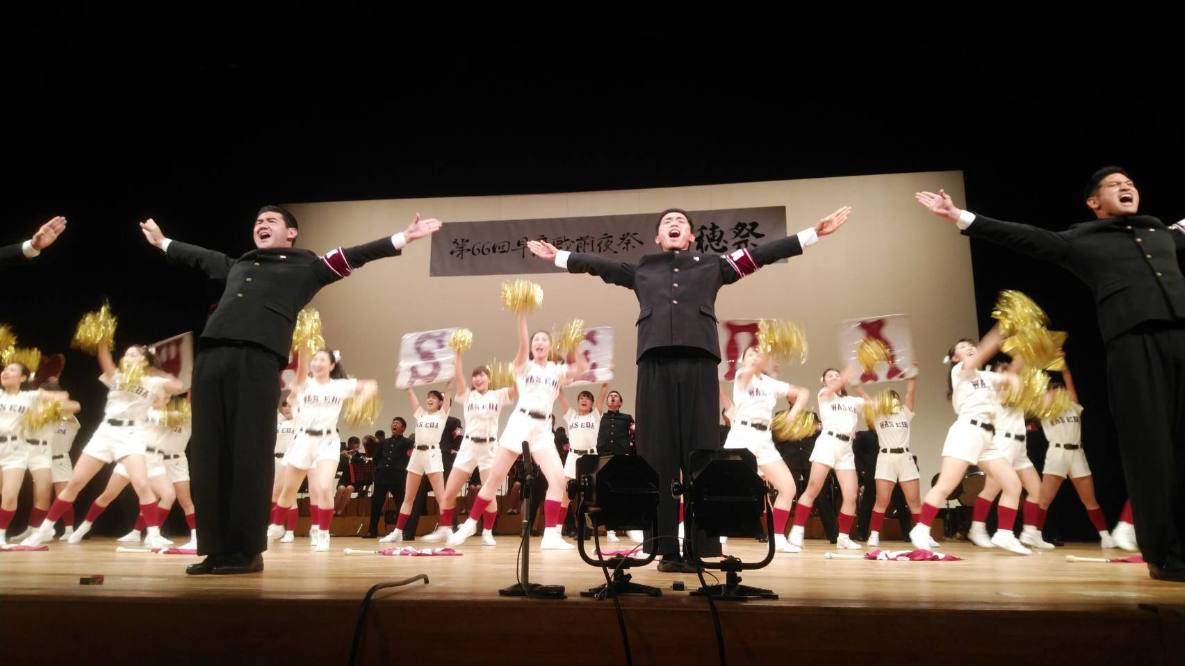 チアリーディングの衣装を着た学生と学生服を着た応援団がステージの上に立ち、両手を広げて応援する様子の写真