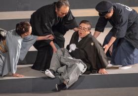 紋付き袴姿の市長が、舞台の段差で尻もちをついており、近くの演者が市長を支えている様子の写真