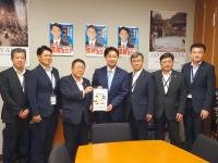 選挙用ポスターが壁に貼られた部屋で、市長を含む男性7名が並んで立ち、市長と男性で要望書を持っている写真