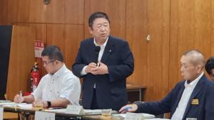 市長が自席で立ちマイクで話をして、両脇に座る男性が資料を見ている写真