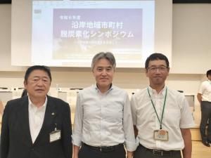「沿岸地域市町村脱炭素化シンポジウム」の文字が映し出されたスクリーンを背景に、市長と参加者2名が並んで撮影した写真