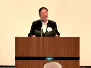 白の胸章をつけた市長が演台で、ファイルを開いて話をしている写真
