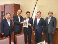 国旗が置かれた机の前で男性2名で要望書を持ち、その横に市長を含む男性3名が並んで撮影した写真