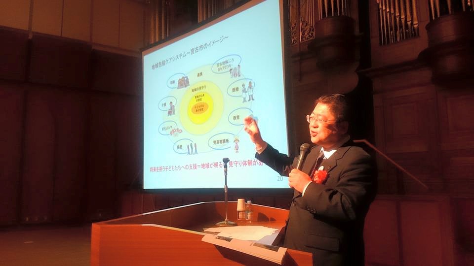 画面が映されたスクリーンの横に置かれた演台で講演を行う様子の市長の写真