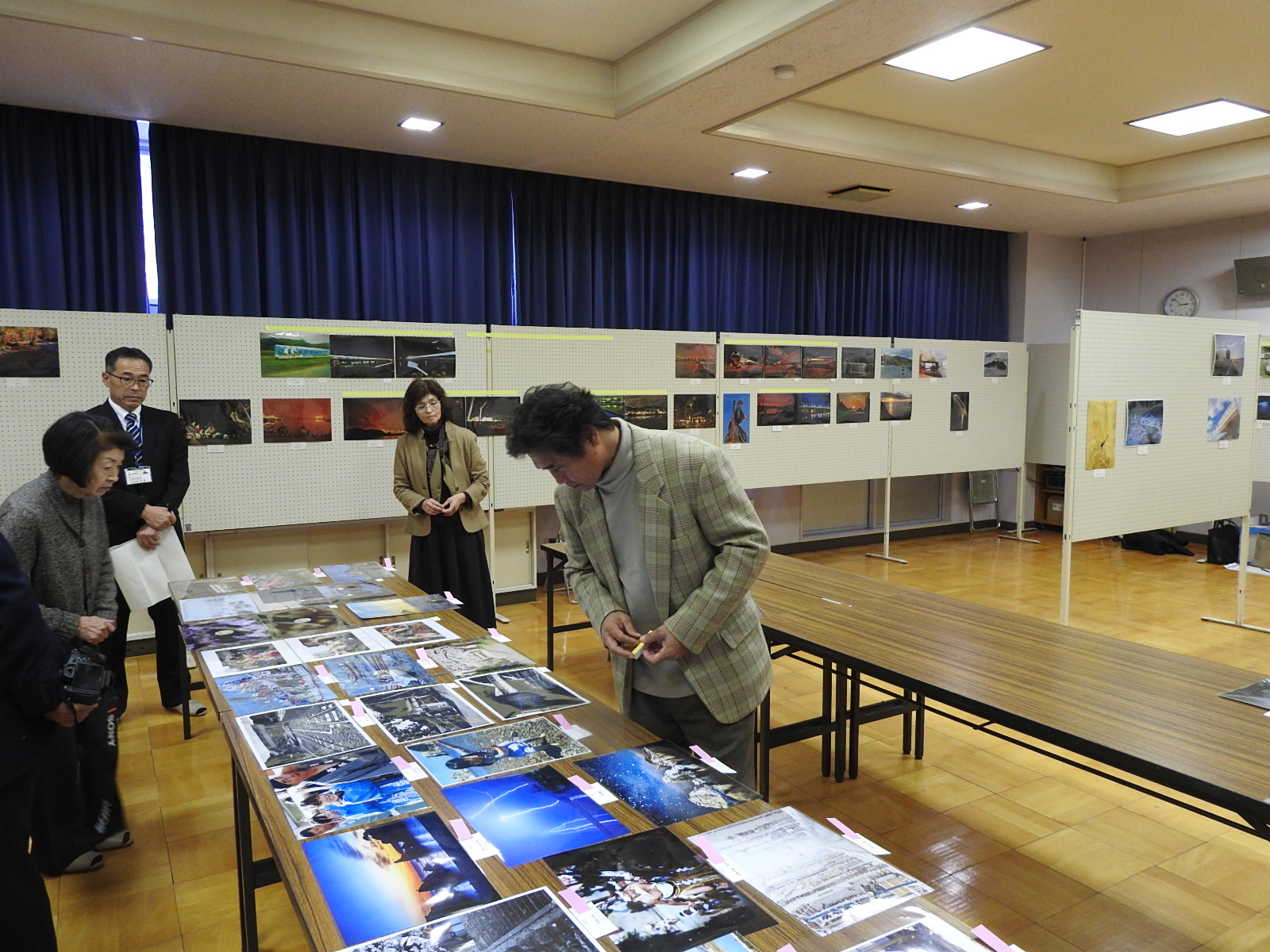 白い展示パネルには二段に写真が展示されており、その手前の長机では2組の男女が立っていて、手前左の女性と手前右の男性が写真を審査している様子を撮影した写真