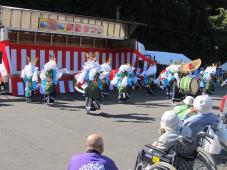 正面紅白の横断幕の前で頭に白くて長いふさふさしたものを被り、衣装をまとって踊っている人たちを車いすの人達が見物している様子を左後方から撮影した写真