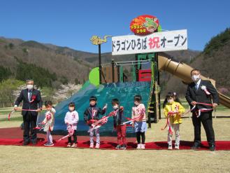 遊具に「ドラゴンひろば祝オープン」と書かれた幕が飾られ、男性2名と6名の子供たちが紅白テープを手に持っている写真