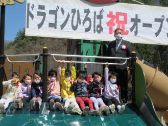 「ドラゴンひろば祝オープン」と書かれた幕が飾られた近くで8名の子供たちが座り、右横に男性が立っている写真