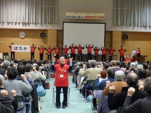 赤いTシャツを着たシルリハおらんど宮古の会の皆さんが前に立って体操をする様子を見ながら参加者達が座りまねをして体操をしている様子の写真