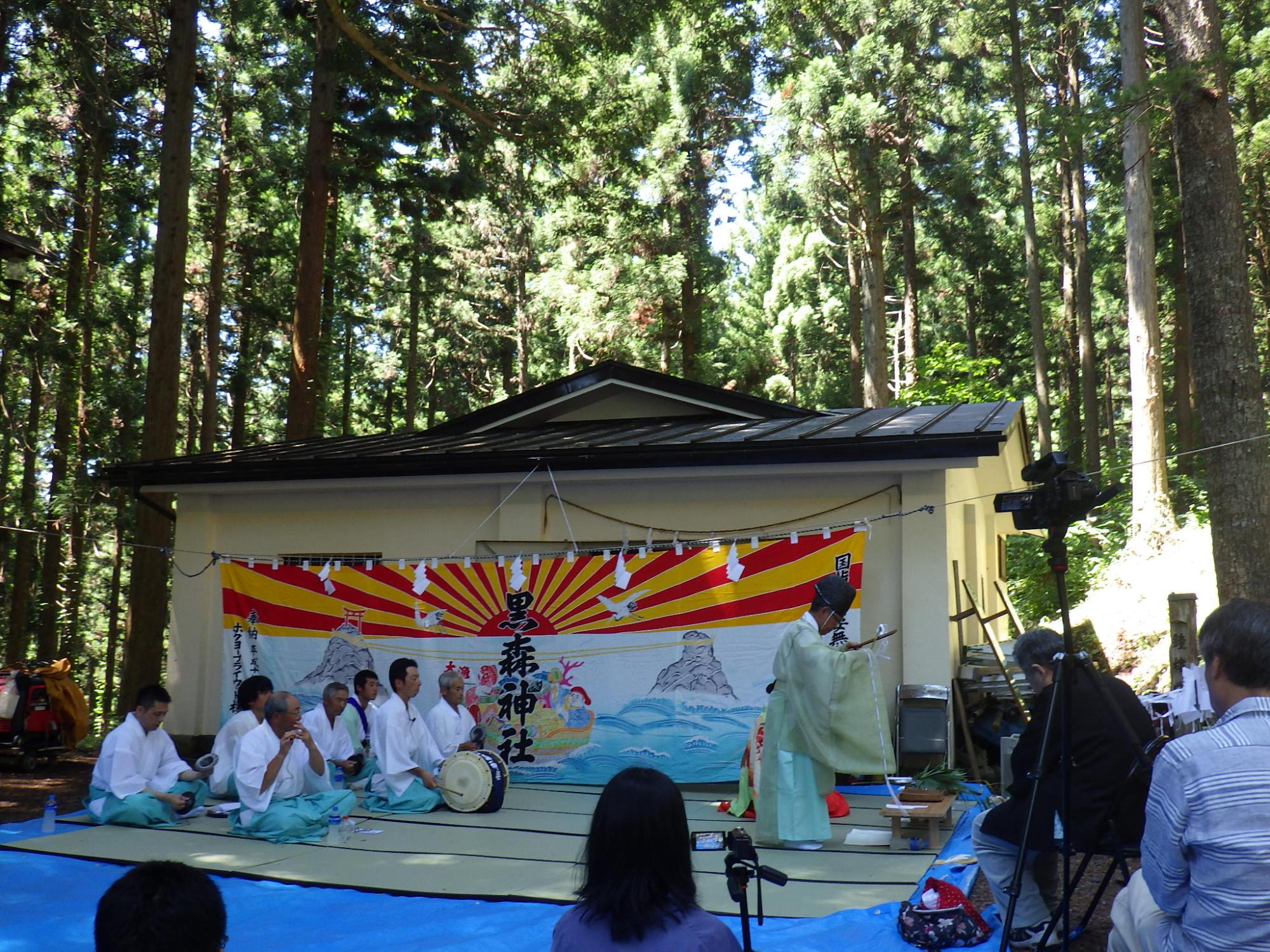 黒森神社と書かれた幕の前で、装束を着た人が立ち、その後ろで水色の袴を着た人が笛や太鼓を演奏している写真