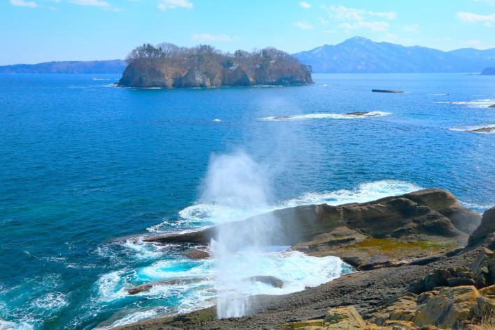 岩の間から海水が吹き出している潮吹穴の奥に見える日出島の写真