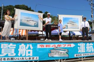 ステージに立ちながら遊覧船の写真を前方に見せる複数の男女を斜め前から写した写真