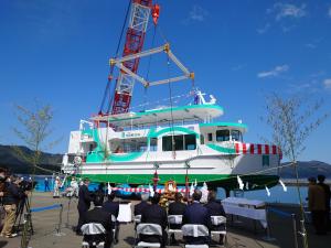 赤色と白色の縦じまの幕が前方につけられた宮古うみねこ丸が、大きなクレーンに吊られて陸地から海に移されている様子を斜め前から写した写真