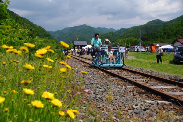 線路の横に黄色の花が咲いており、沢山の参加者が見守る中、水色のレールバイクに乗った3名が走っている様子の写真